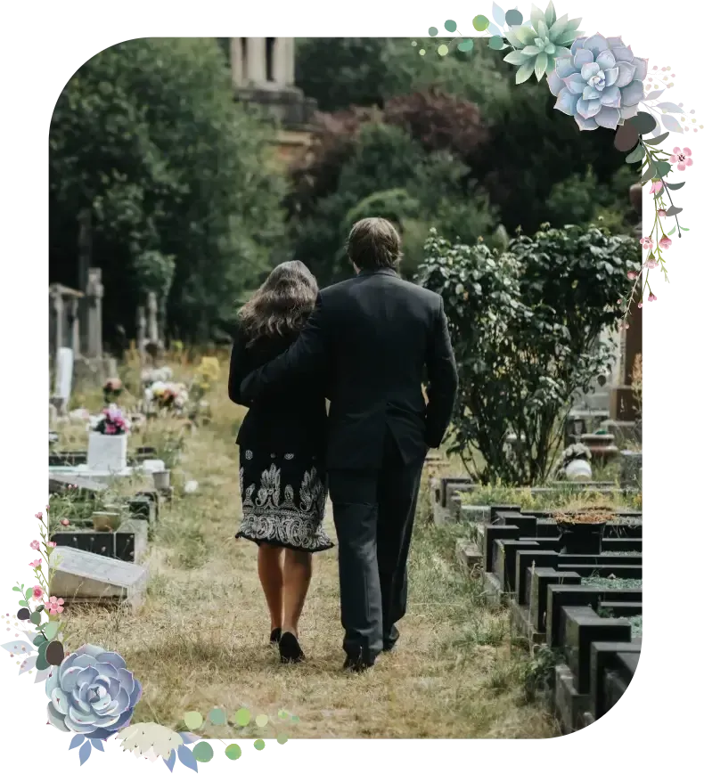 Two people walking arm in arm down a cemetery path, bordered by gravestones and flowers.