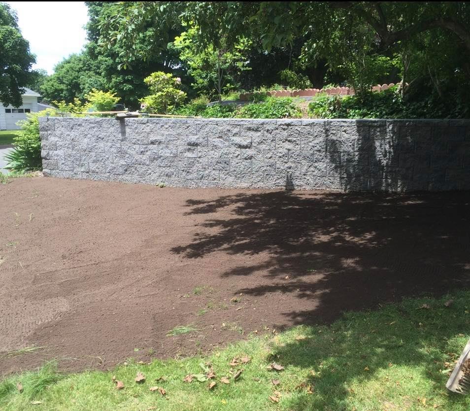 A large, gray stone retaining wall separates a freshly tilled brown area from green grass.
