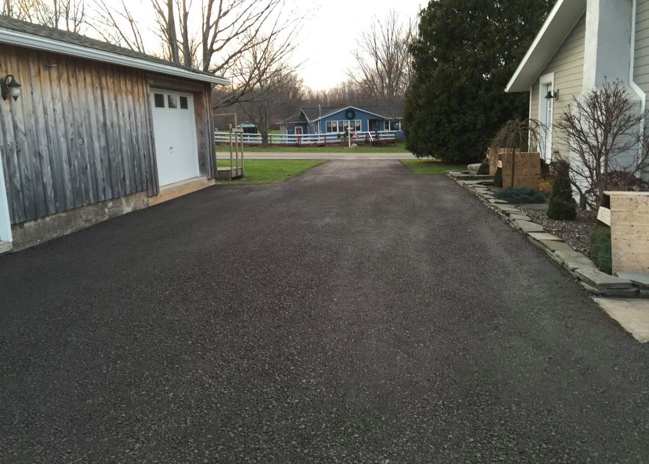 Asphalt driveway leading past a wooden garage and a beige house, grassy lawn in background.