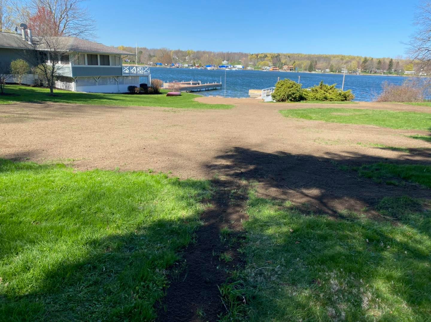Lawn leading to a lake. A house with a dock is visible. Brown earth patch in the middle, blue sky and water.