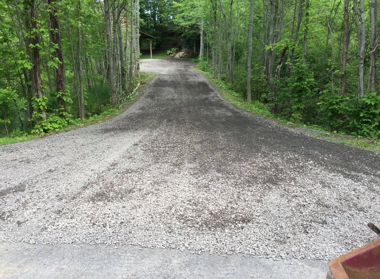 Gravel driveway curves through a wooded area. Light gray gravel contrasted with dark soil.