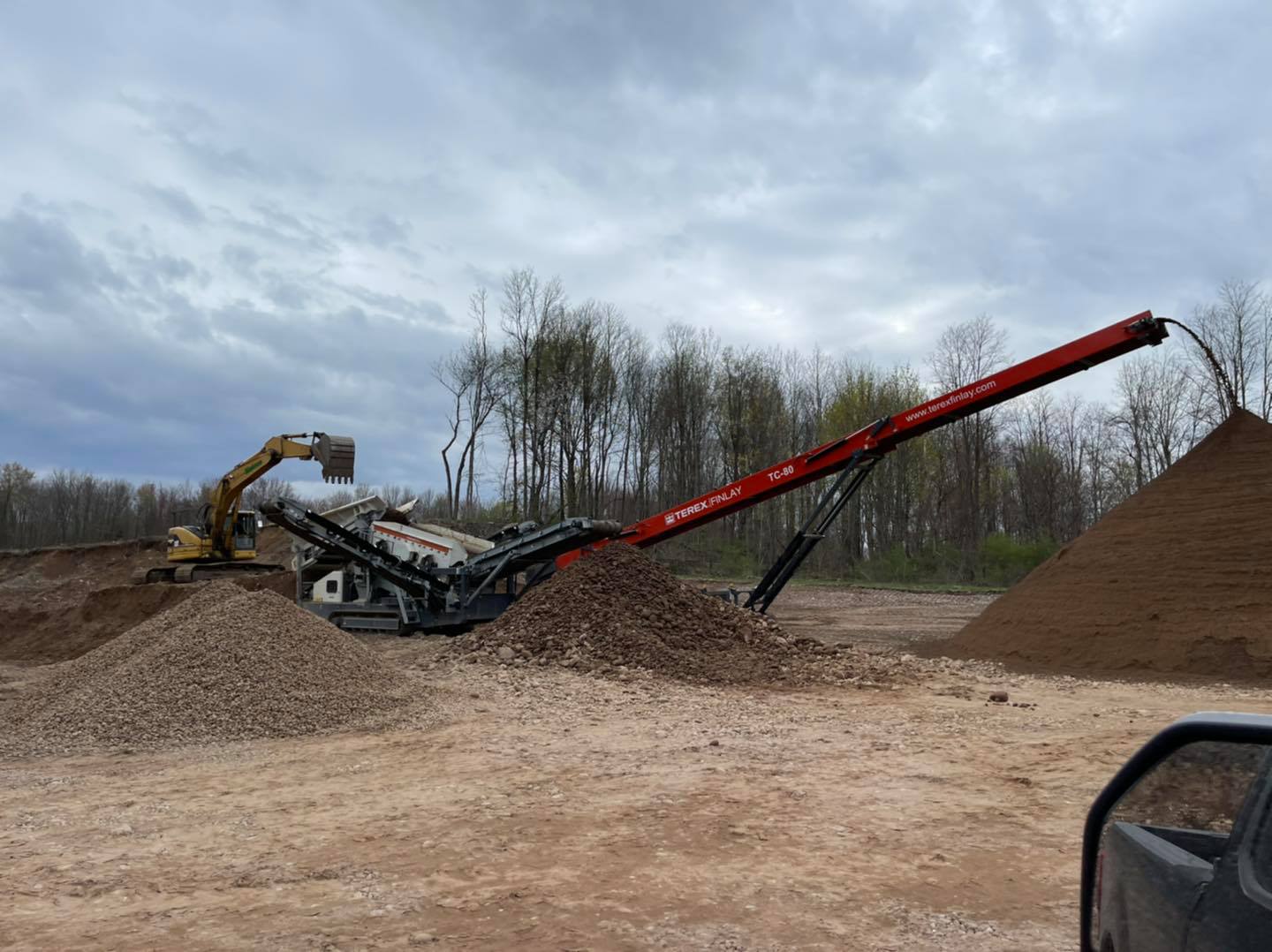 An excavator loads rocks into a rock-crushing machine, which then deposits crushed rock onto piles. Outdoors, cloudy sky.