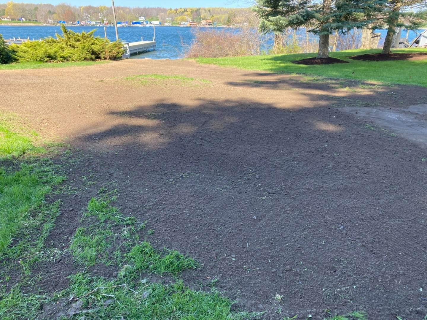 Newly tilled soil ready for planting, near a lake with a boat dock and trees. Sunny day.