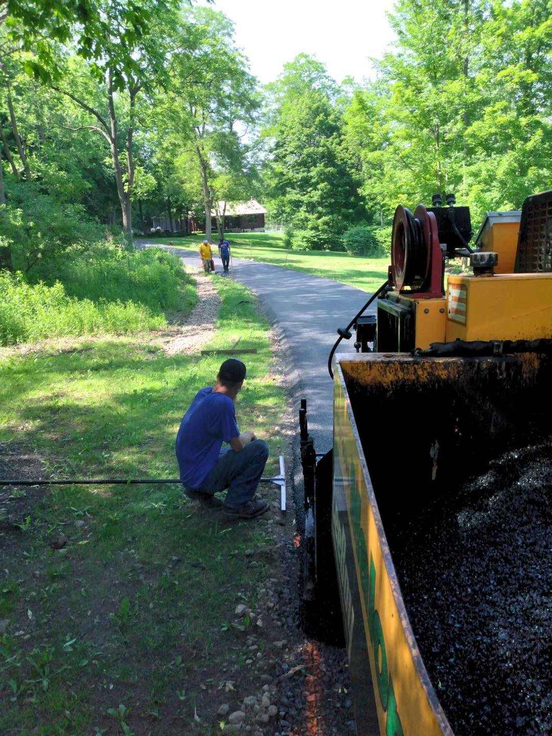A worker kneels near a yellow asphalt paving machine, laying asphalt on a path near trees.