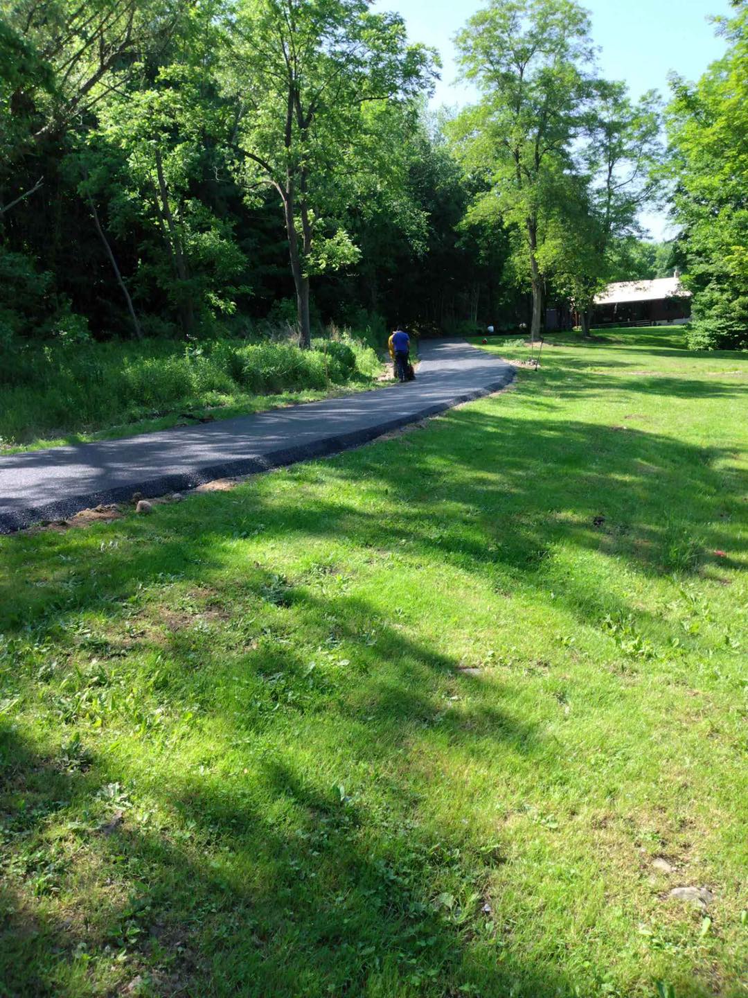 Paved path winds through green grass and trees; a person pushes a lawnmower on the path.