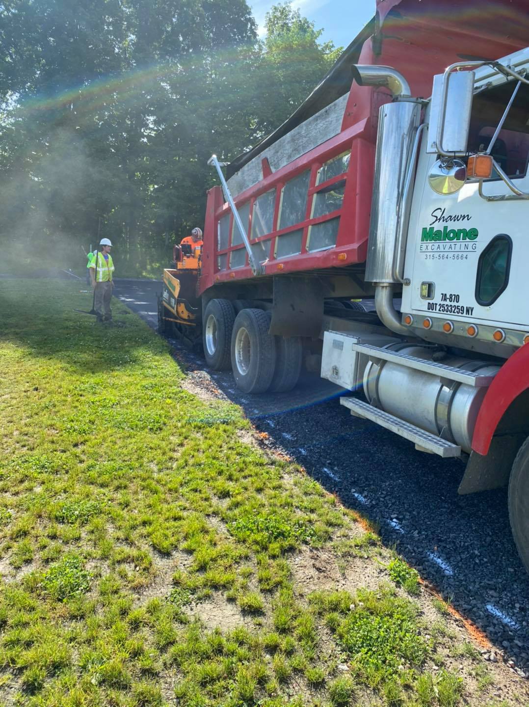 Dump truck unloading asphalt onto a roadway, with workers in safety vests and a green grassy area.