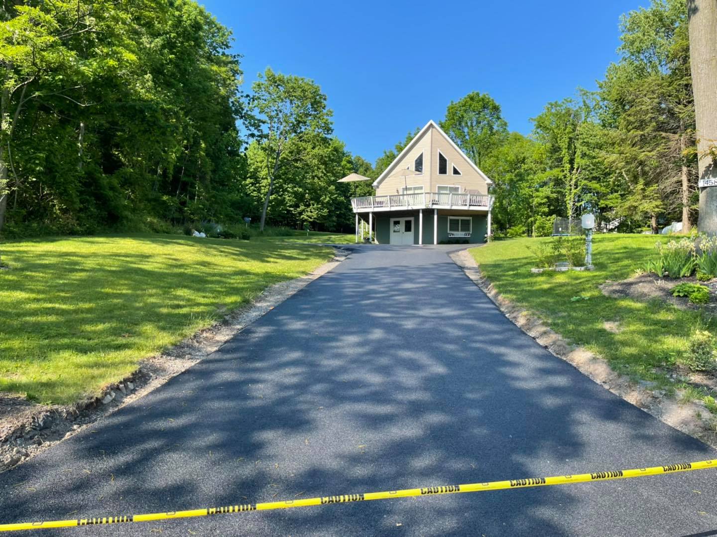 Black asphalt driveway leading to a two-story house with a green porch, surrounded by green grass and trees.