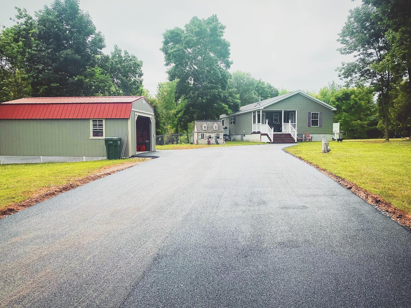 Asphalt driveway leading to a green house with a porch and a red-roofed shed on a grassy lot.
