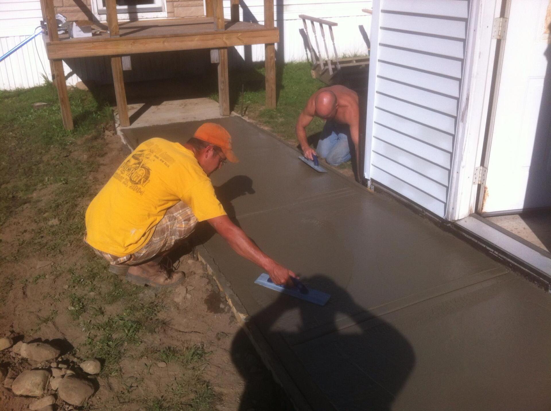 Two people smoothing wet concrete for a sidewalk next to a building. One in a yellow shirt, the other shirtless.