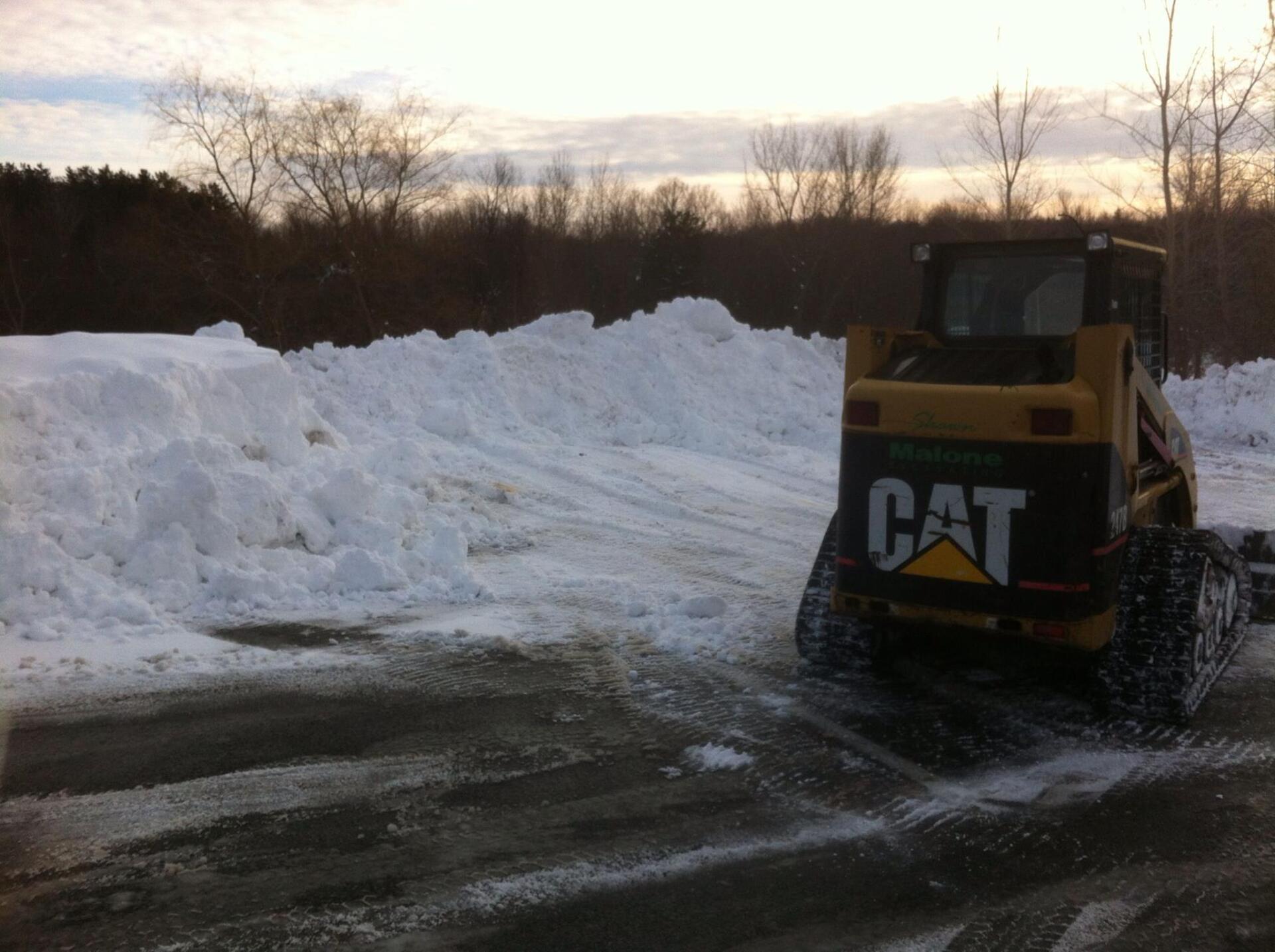 Yellow CAT skid steer removing snow, clearing a paved area. Snowbank in the background.