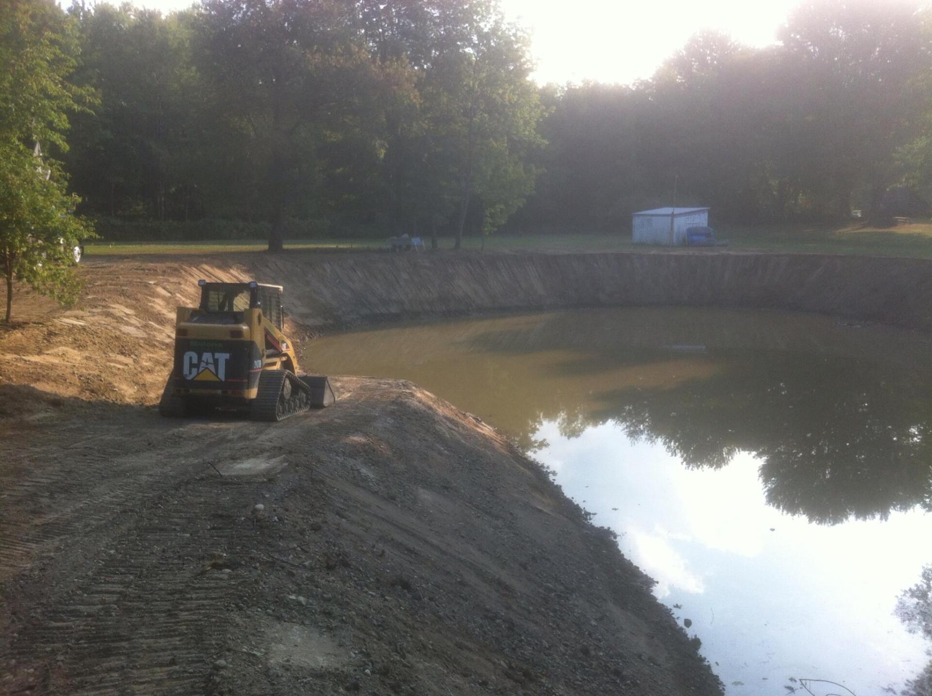 A small CAT skid steer loader near the edge of a newly dug pond. Trees and a white shed are in the background.