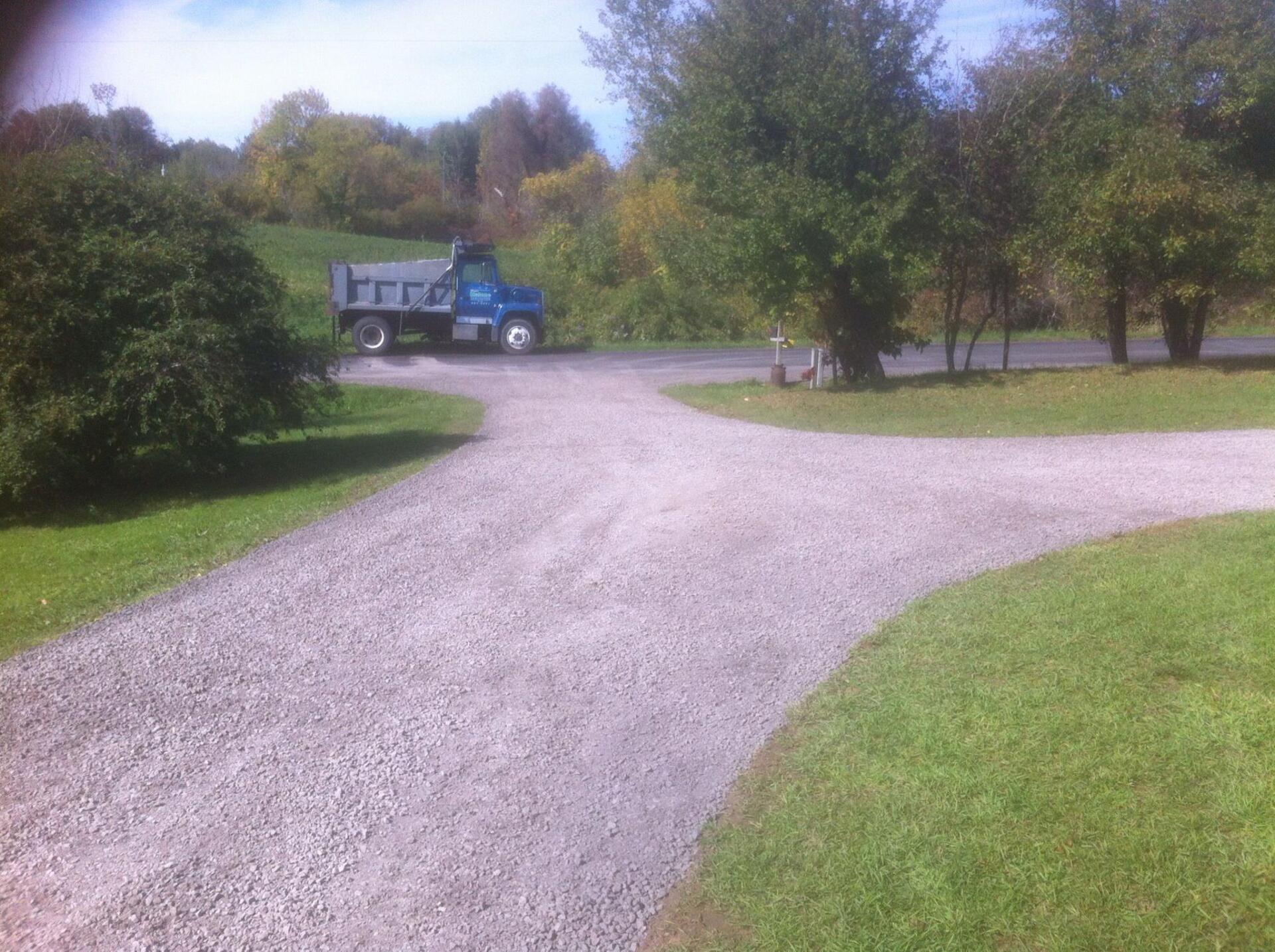 Gravel driveway with a blue dump truck parked near trees and green grass.