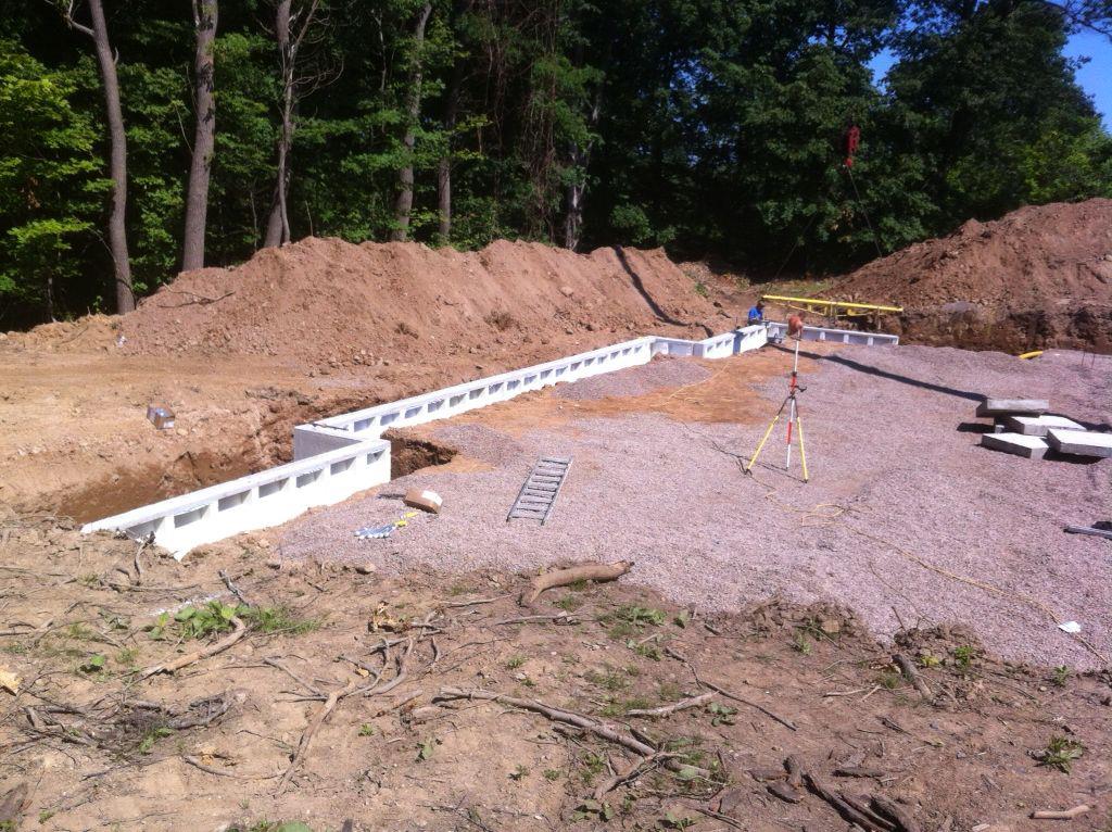 Construction site: white cinder block foundation walls, gravel base, earth mounds, trees in background.
