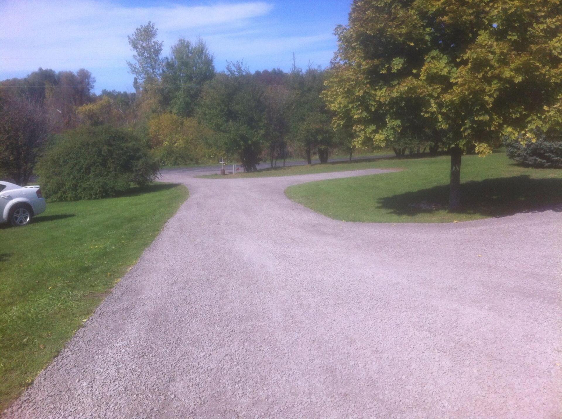 Gravel driveway splits into two directions, bordered by green grass and trees under a blue sky.