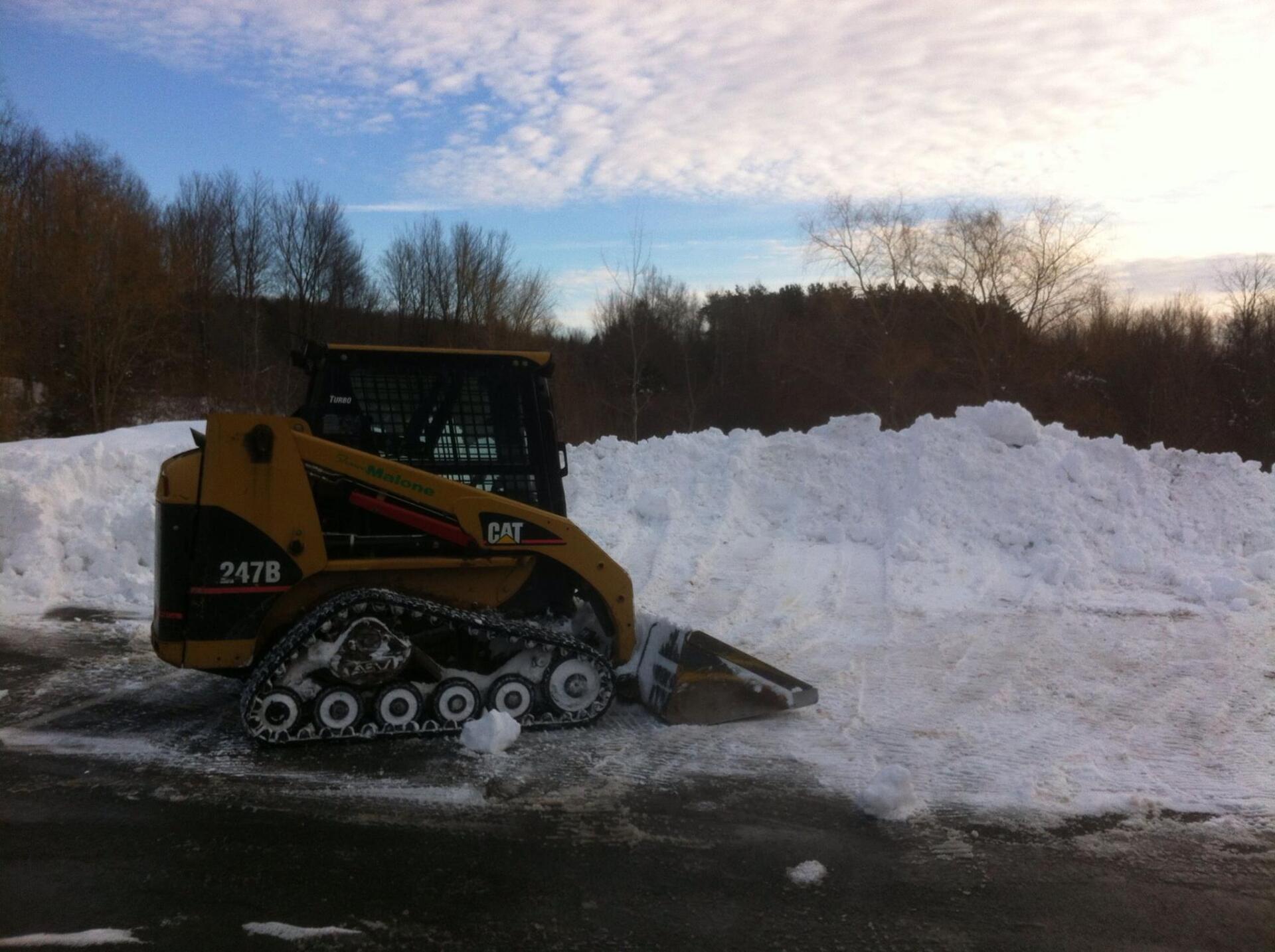 Yellow CAT 247B skid steer clearing snow from a parking lot on a cloudy day.