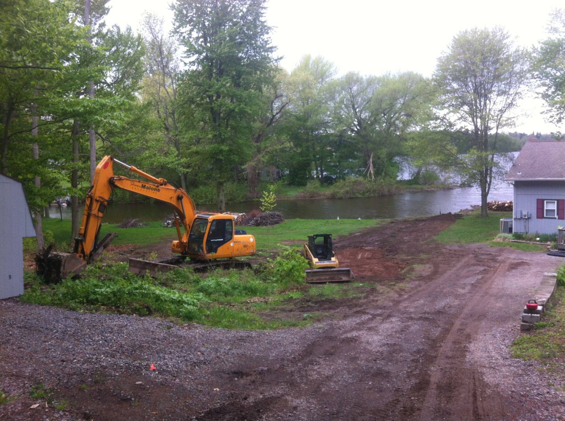 Yellow excavator and small machine clearing land near a body of water, next to trees and a house.