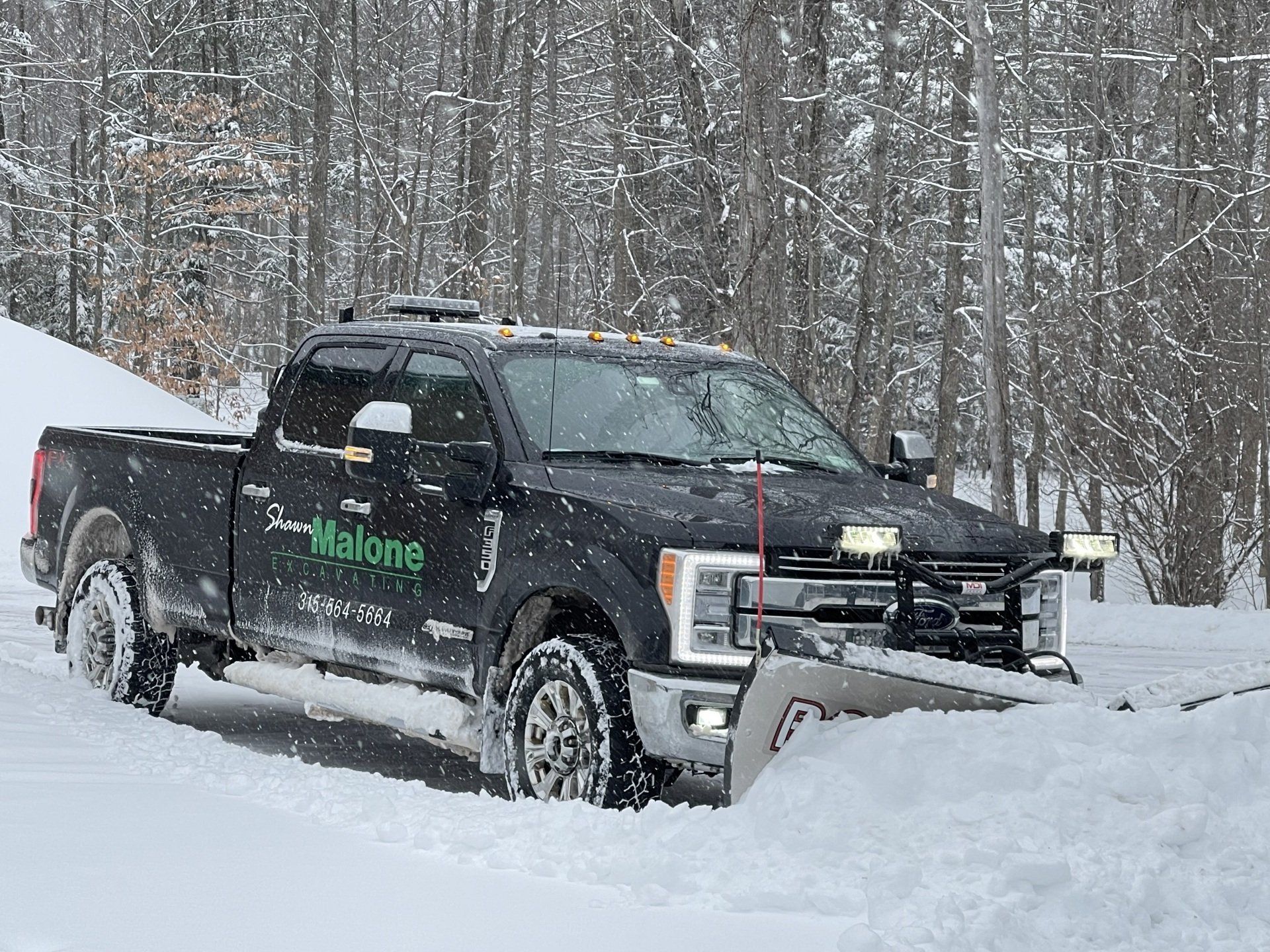 Black pickup truck plowing snow on a snowy road with trees in the background.