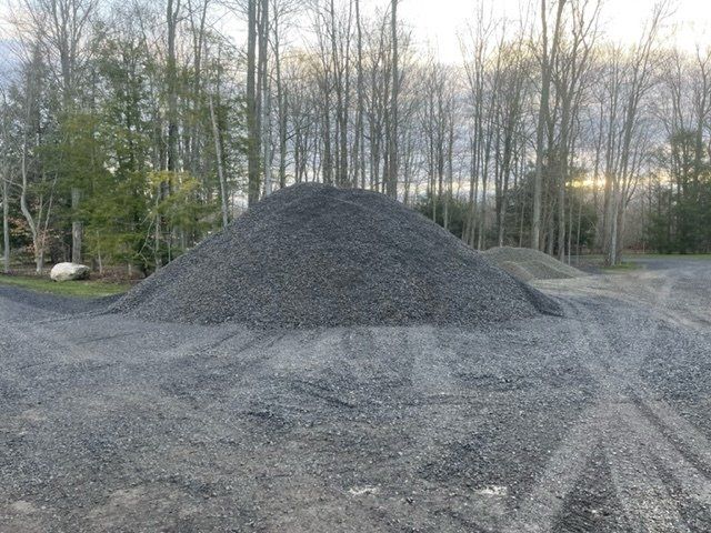 Large pile of dark gravel in an outdoor setting with trees in the background.