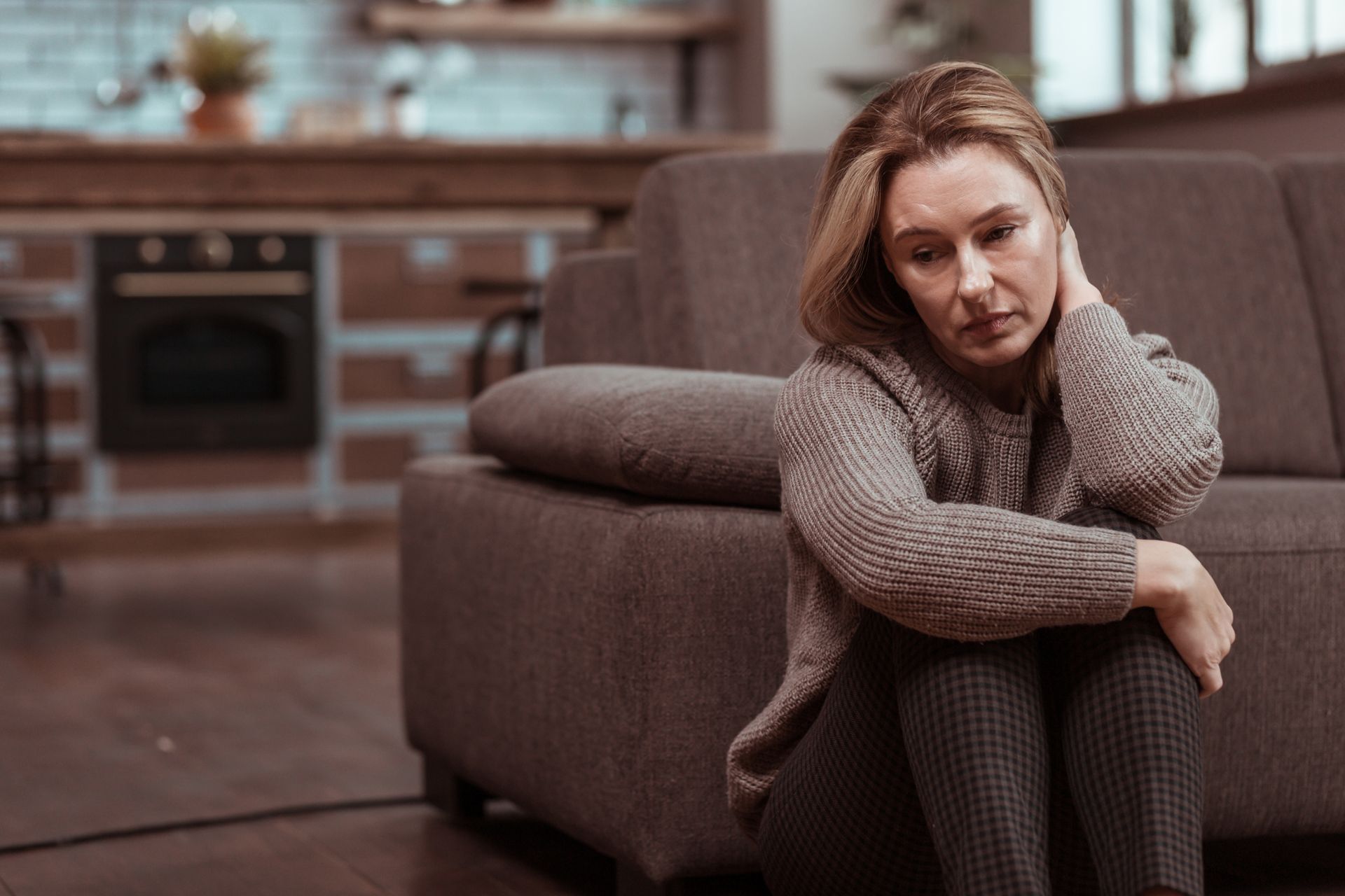 Woman sitting on the floor beside a sofa, resting her head on her hand, in a kitchen living room