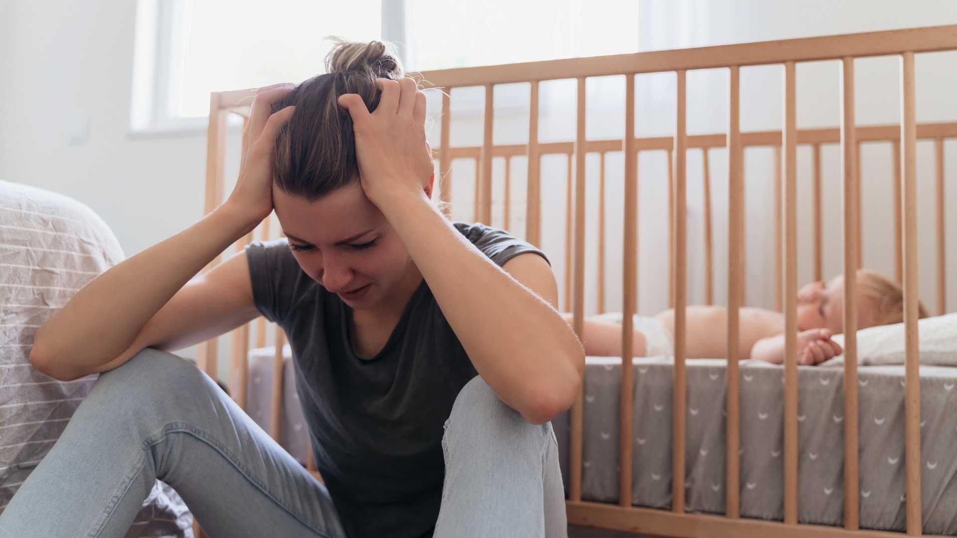 Stressed parent sitting on the floor beside a crib, holding their head while a baby sleeps inside.