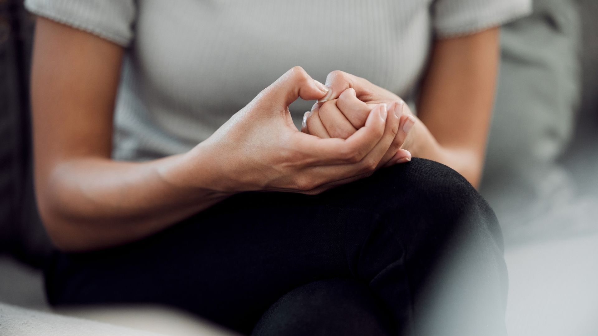 Hands clasped in lap while seated, wearing a gray shirt and black pants.
