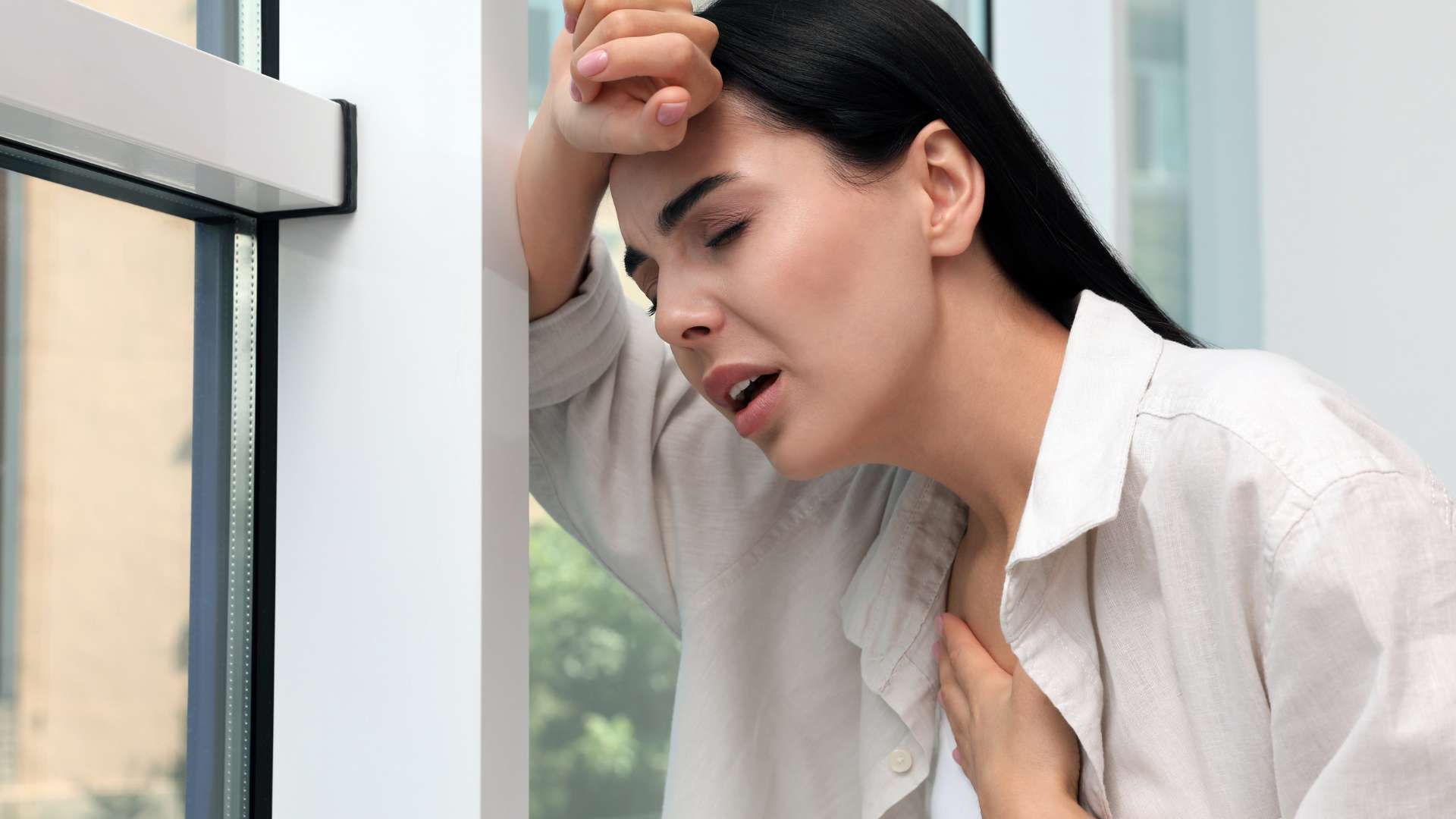Woman in a white shirt leaning against a window, holding her forehead with a strained expression.