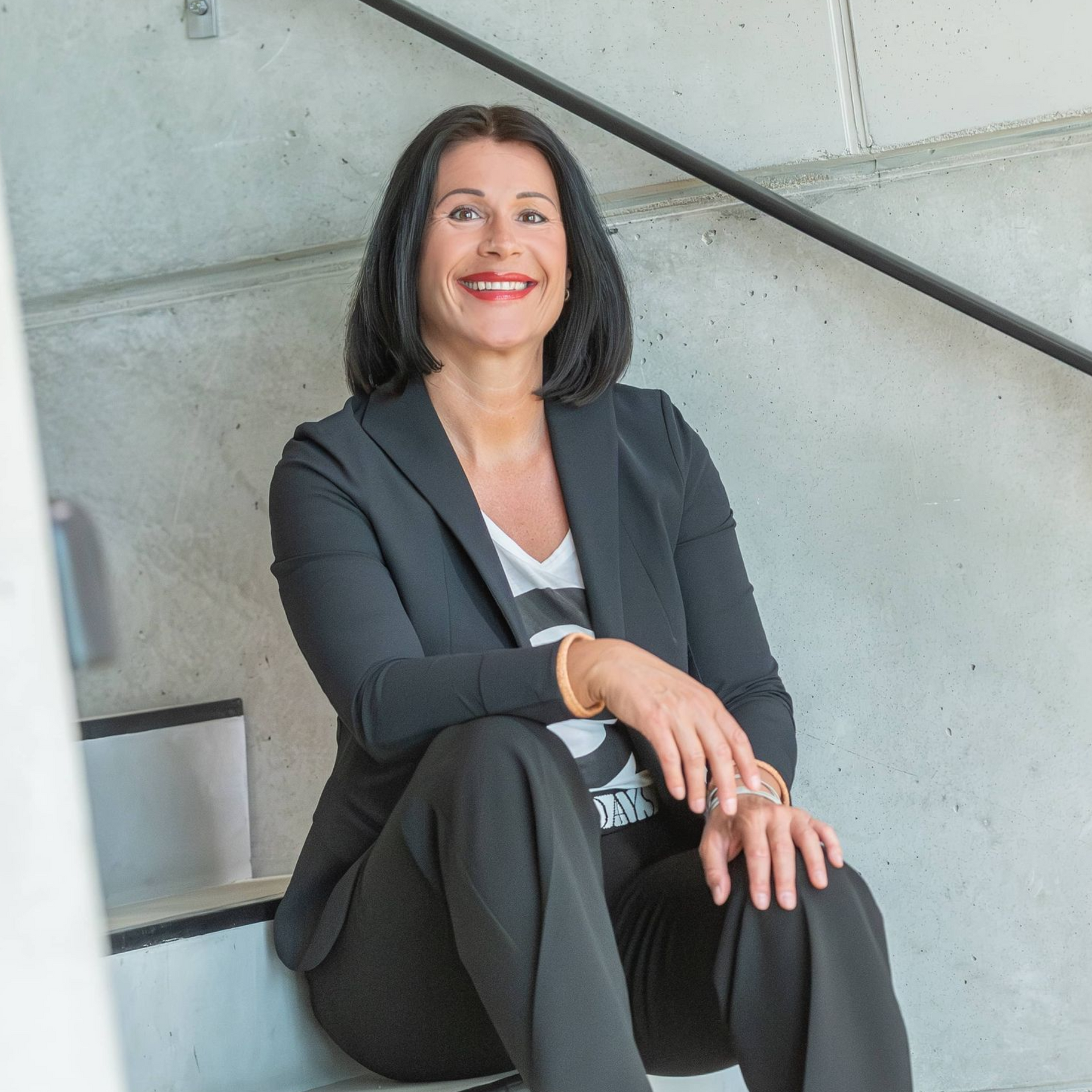 Eine Frau in einem dunklen Blazer sitzt lächelnd auf einer Treppe in einem modernen Betoninterieur.