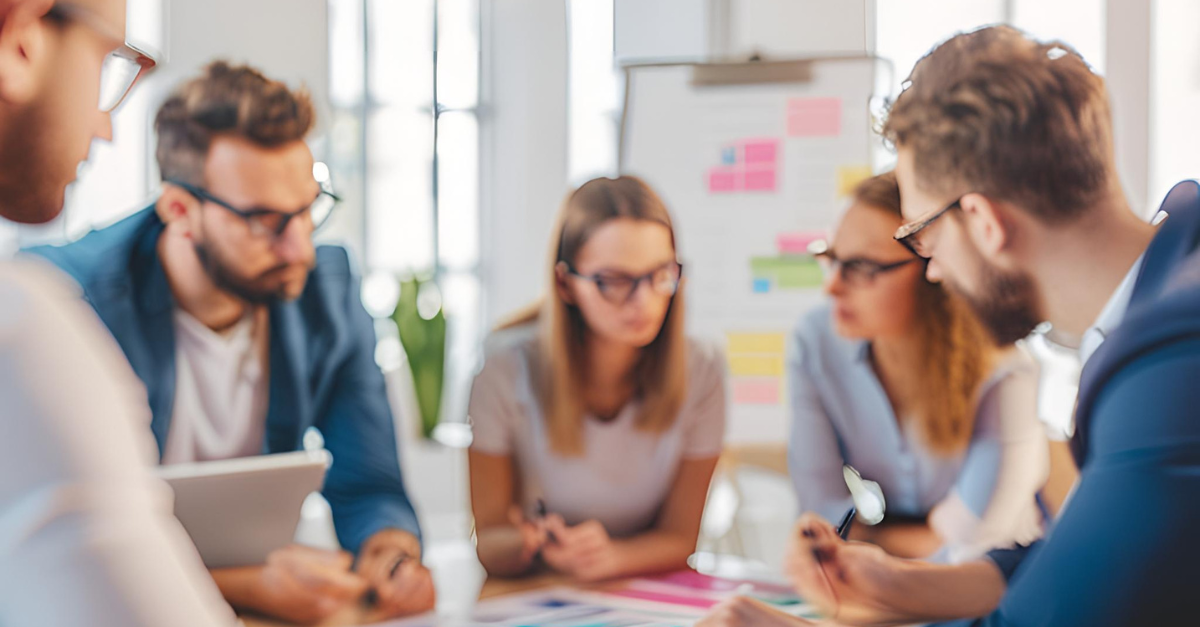 Group of people in glasses at a table, discussing and collaborating, with a whiteboard in the background.