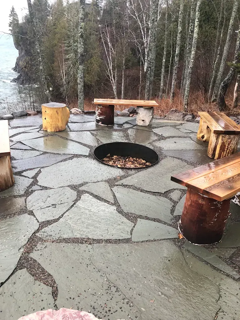 Outdoor stone patio with a circular fire pit and wooden benches beside a snowy forest