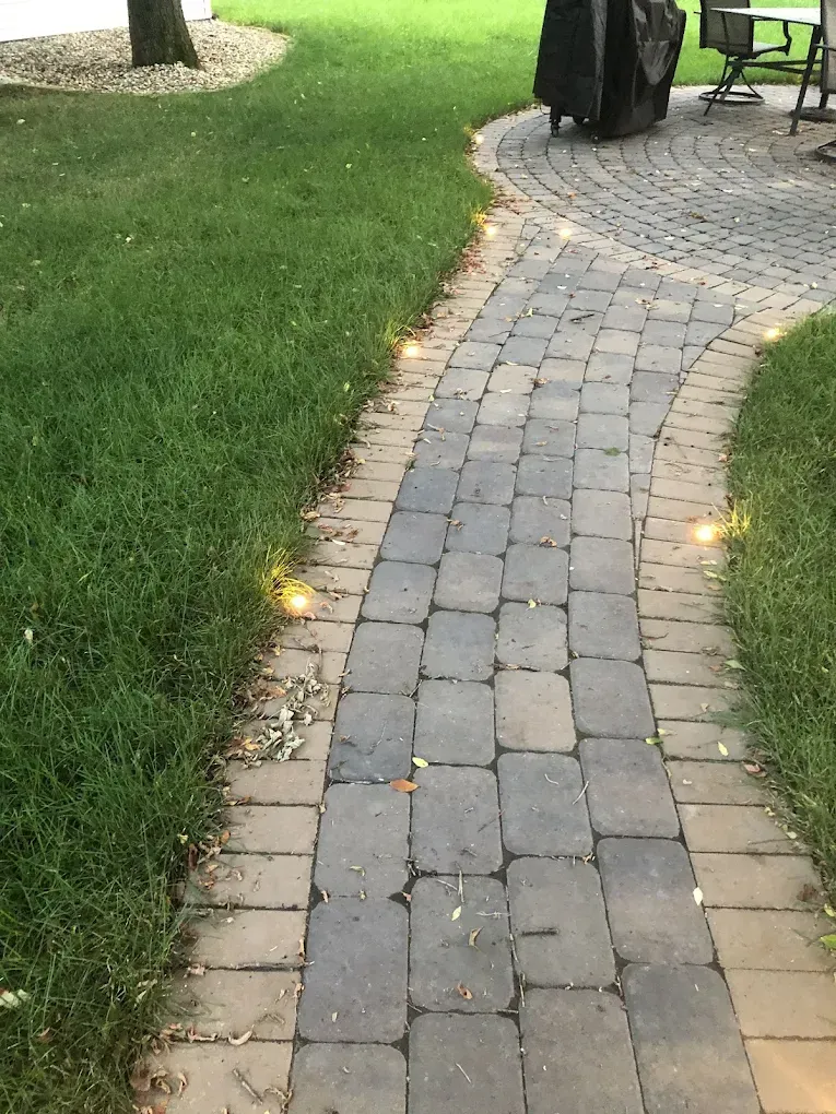 Curved gray stone walkway beside green grass, lined with small ground lights near a patio area.