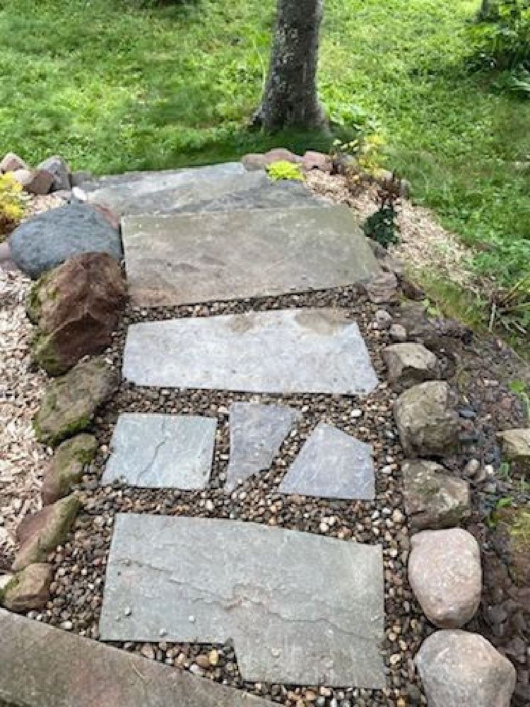 Stepping-stone path of flat stone slabs through a grassy garden, bordered by rocks and a tree trunk.