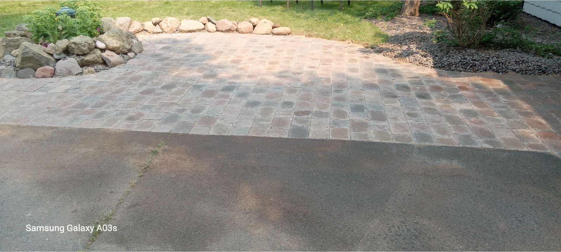 Brick driveway beside dark asphalt, bordered by rocks and green landscaping.