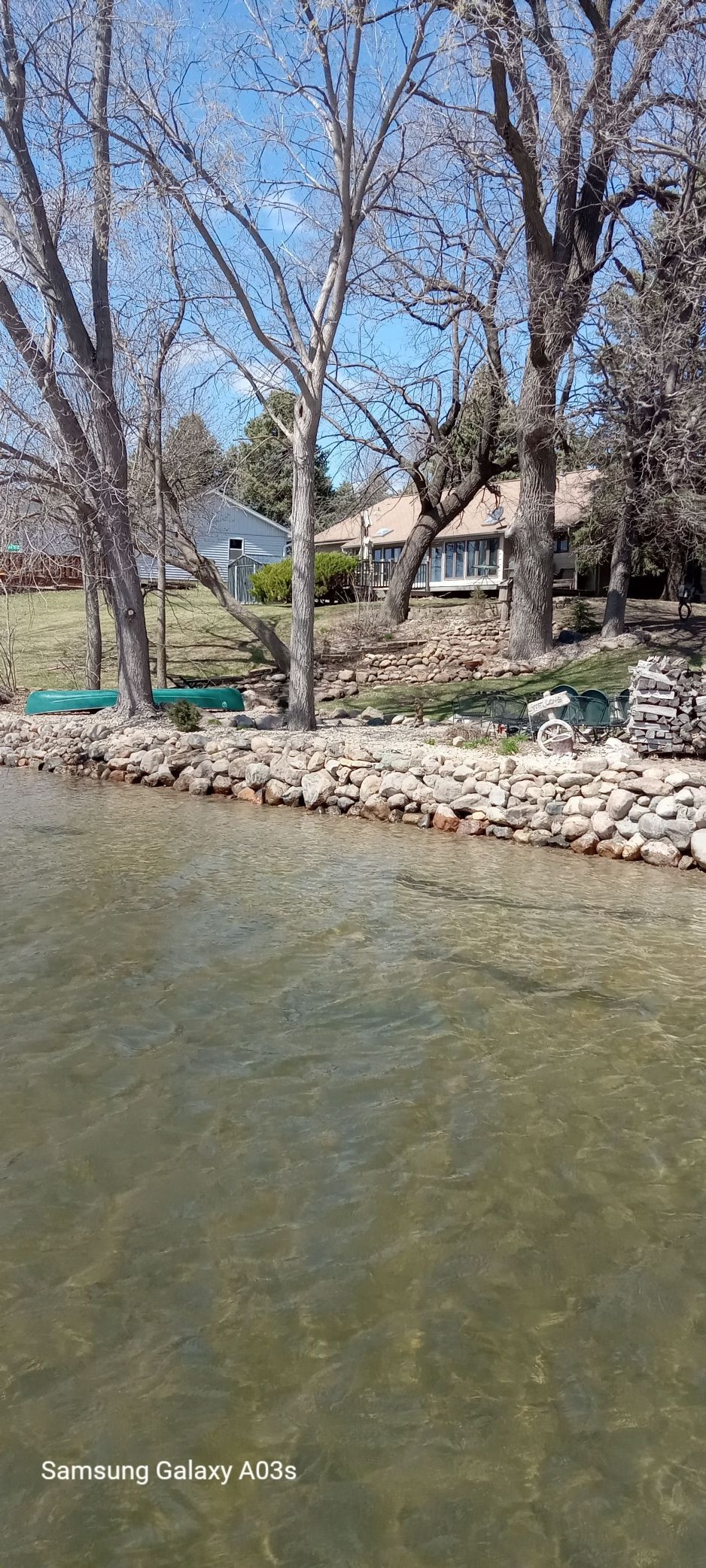 Rocky lakeshore with green water, leafless trees, and houses in the background