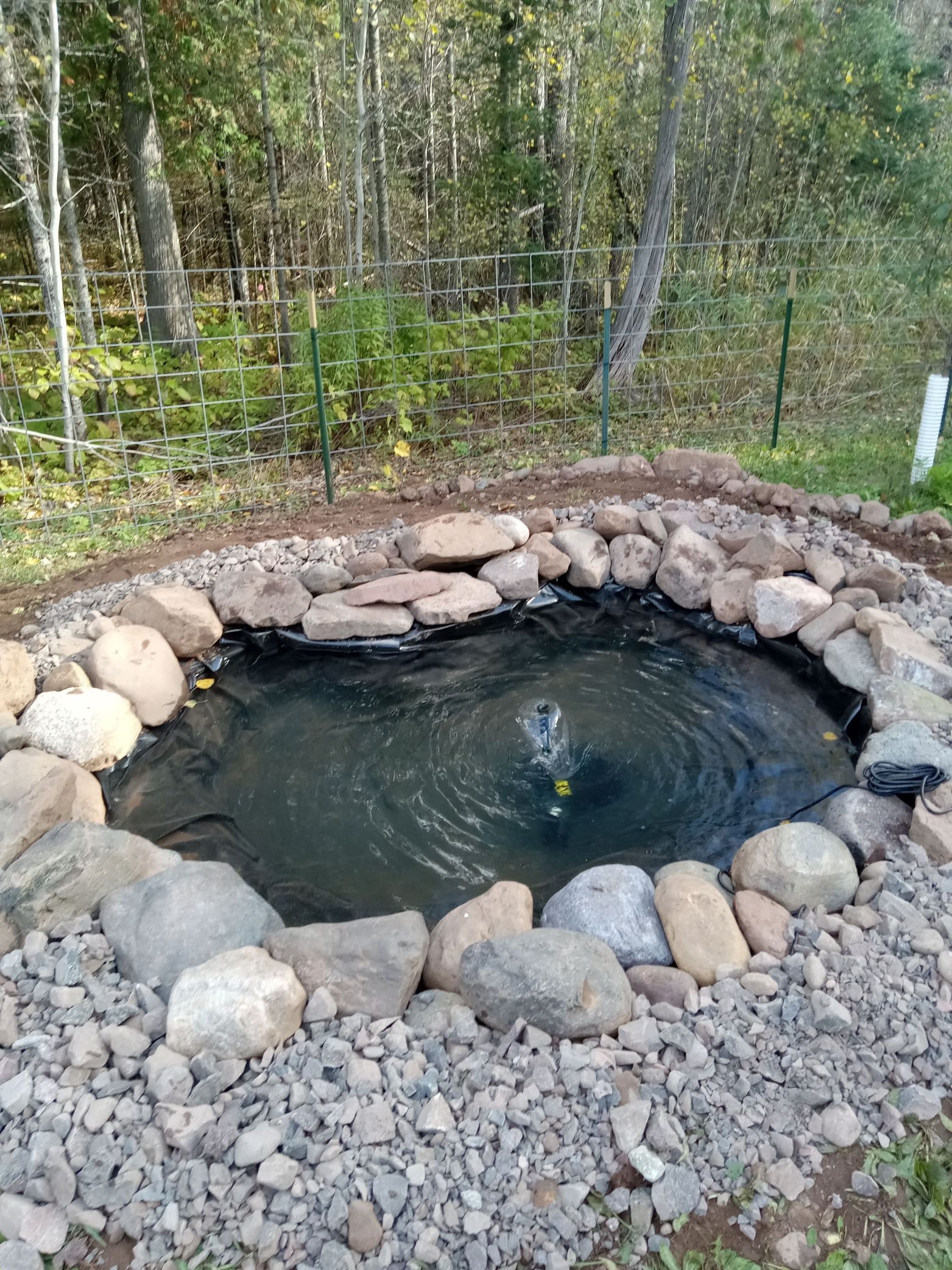 Small stone-lined pond or spring with dark water, set in a wooded area with gravel and rocks.