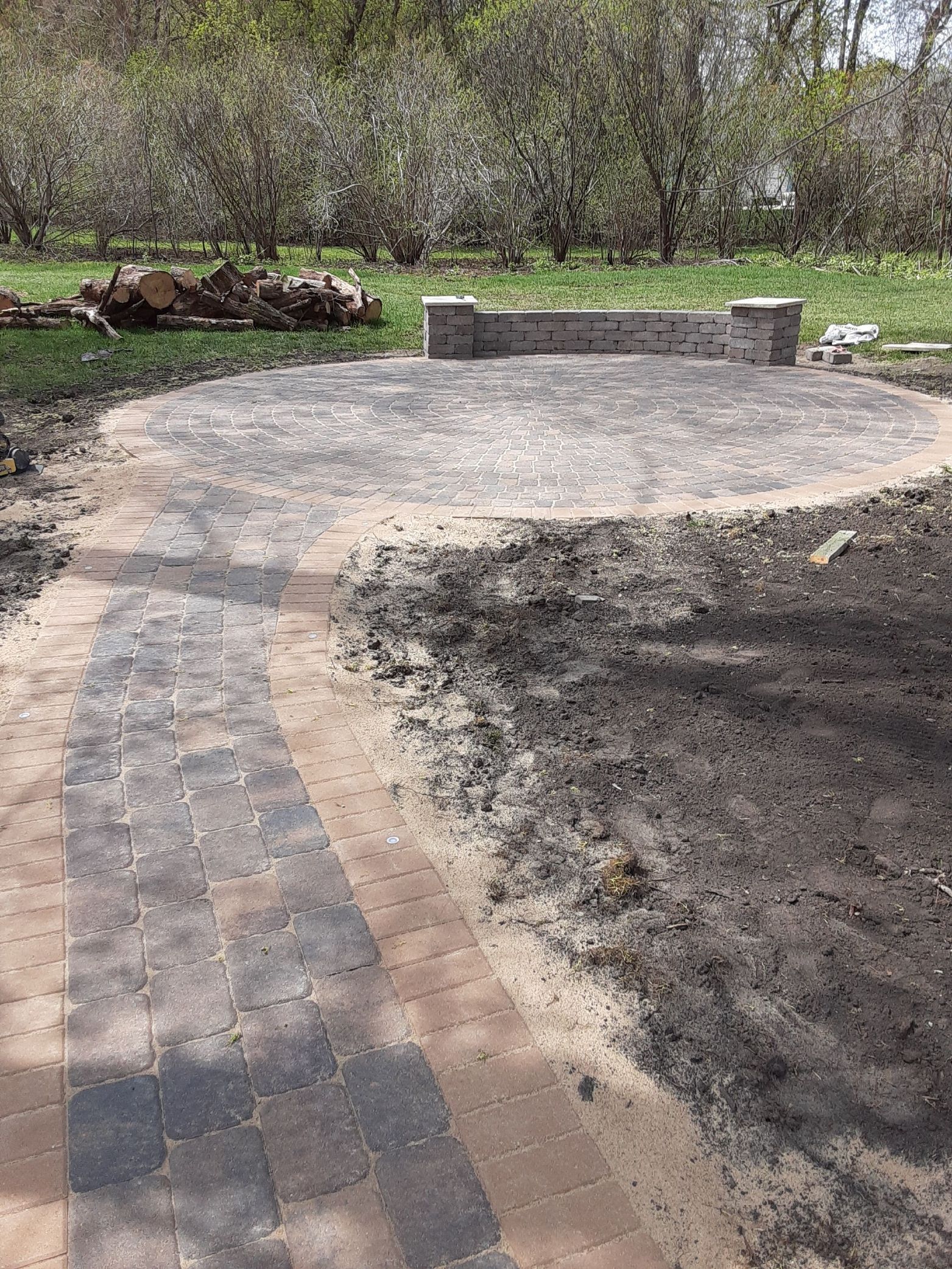 Curved brick path leads to a circular stone patio with a small bench in a wooded park.