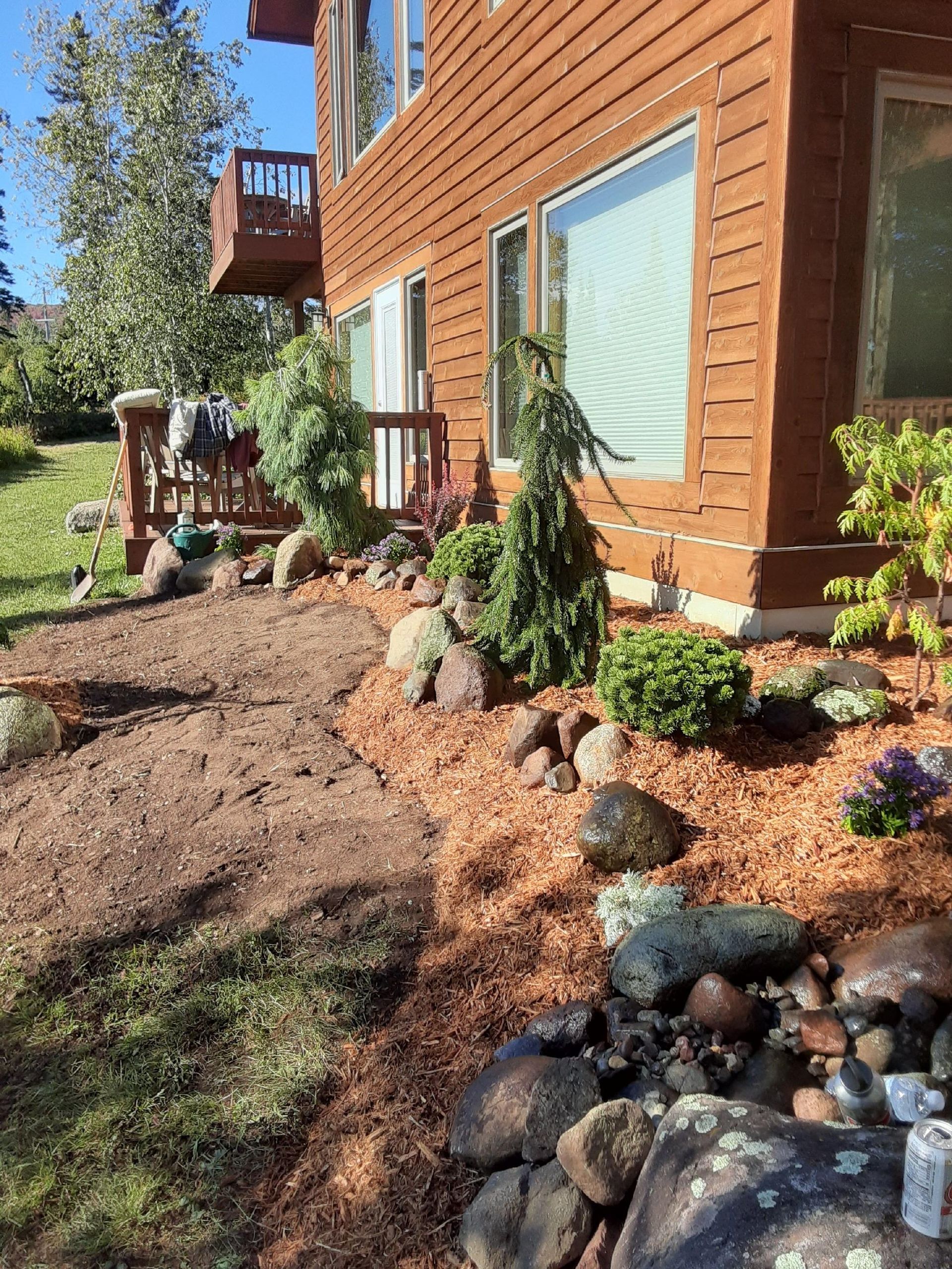 Sunlit wooden house with landscaped garden, mulch beds, rocks, and small plants beside a patio.