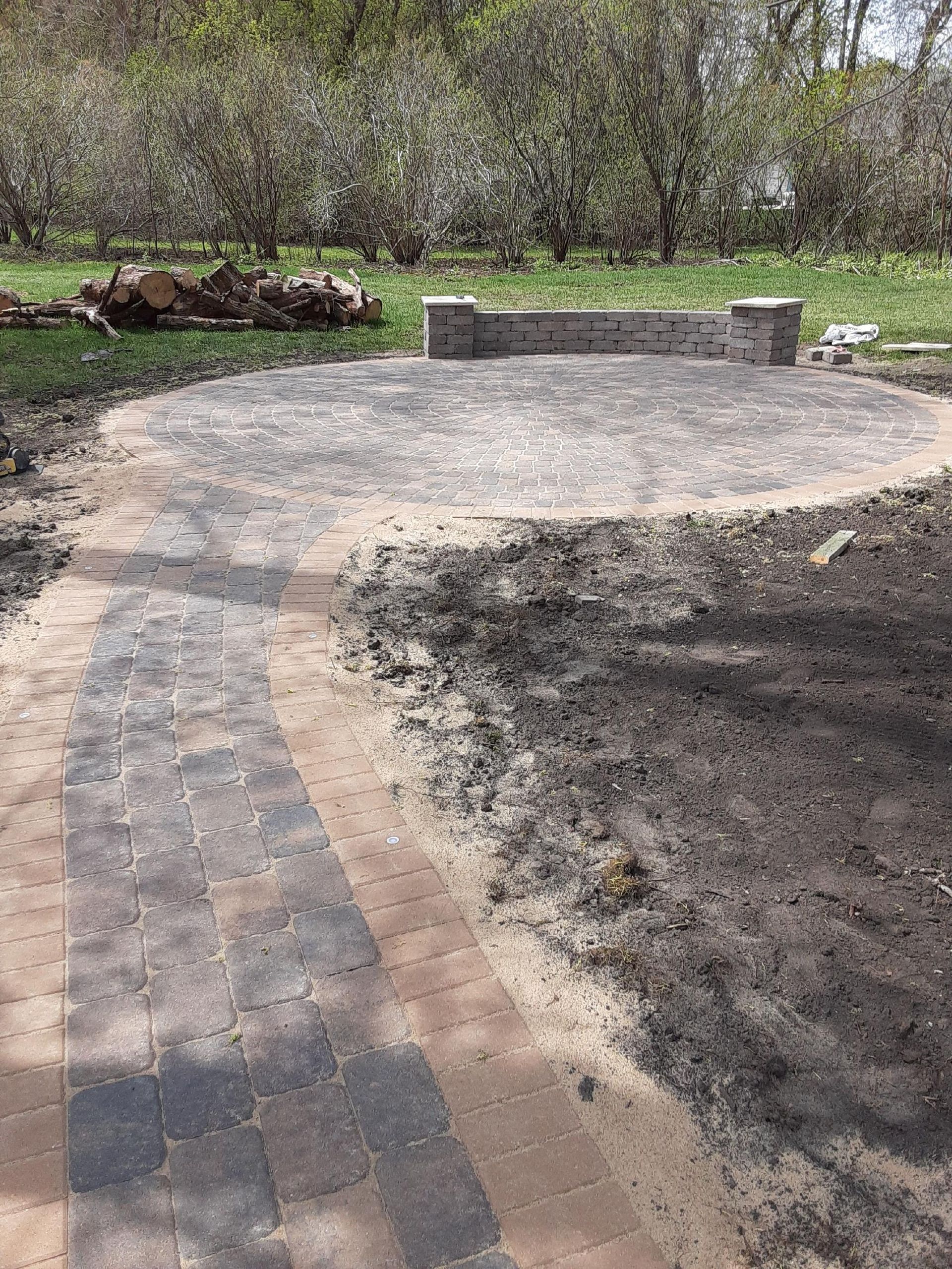 Stone-paved path leading to a circular patio with a low brick wall in a wooded park setting