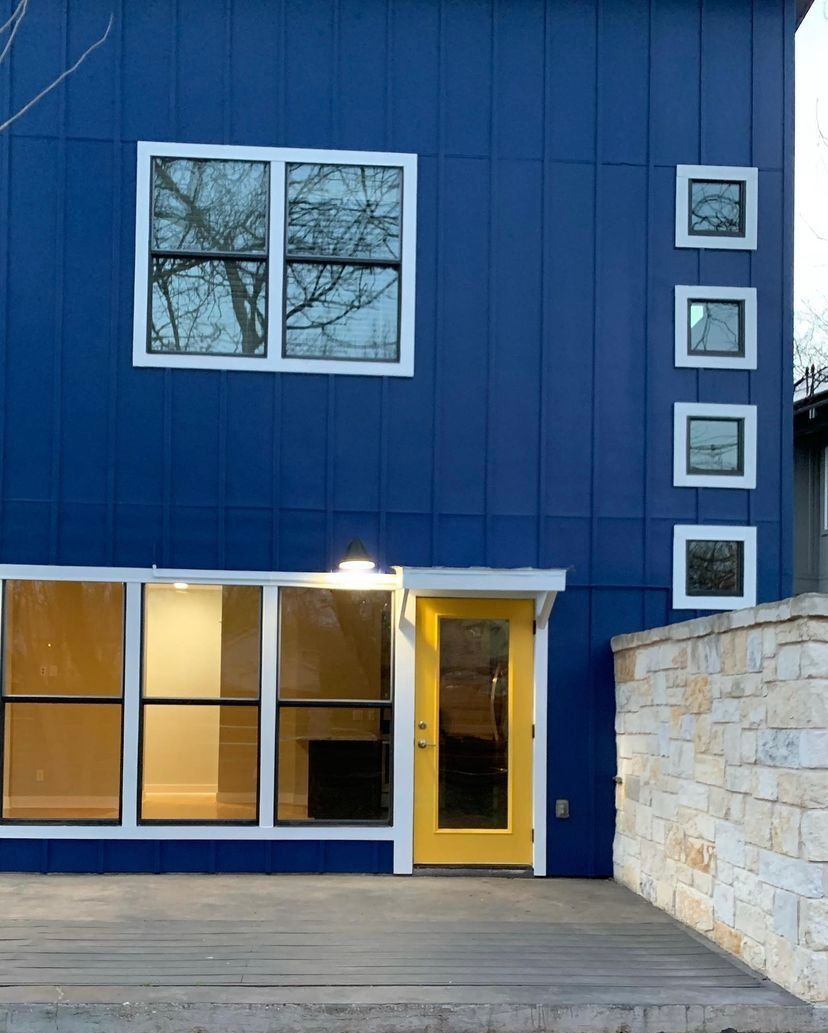 Blue modern house facade with large windows, a yellow door, and a stone wall beside a wooden deck