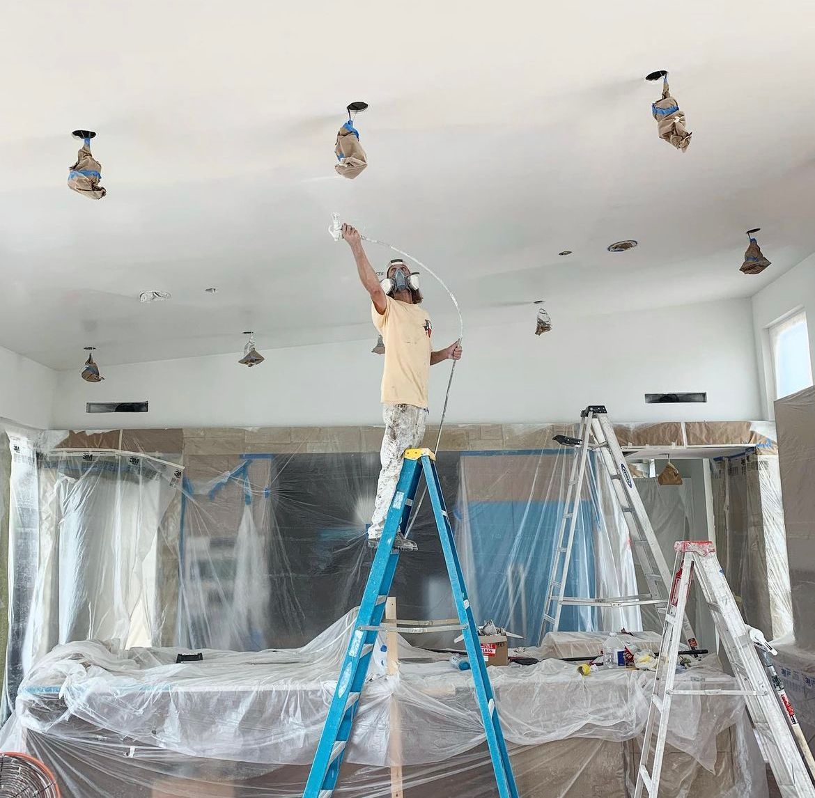 Worker on a ladder painting a ceiling in a room covered with plastic sheeting.