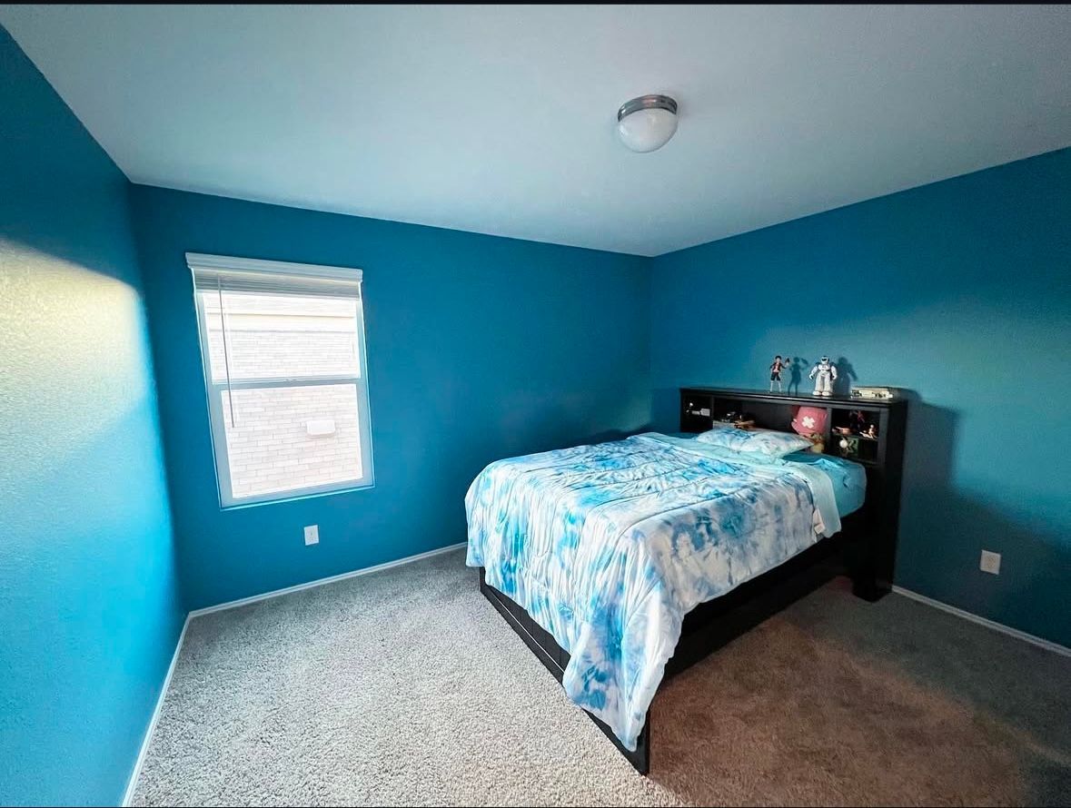 Bright blue bedroom with a bed, dresser, window, and carpeted floor.