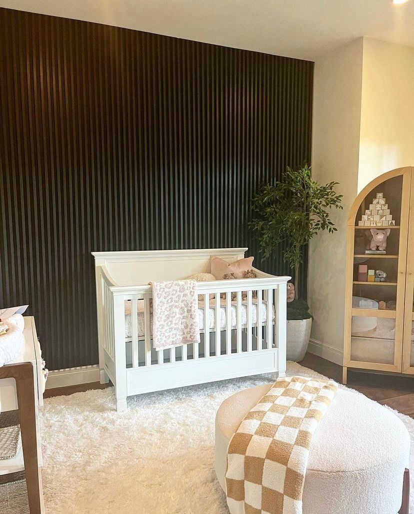 White crib in a beige nursery with dark wood accent wall, plush rug, and arched shelf with decor.
