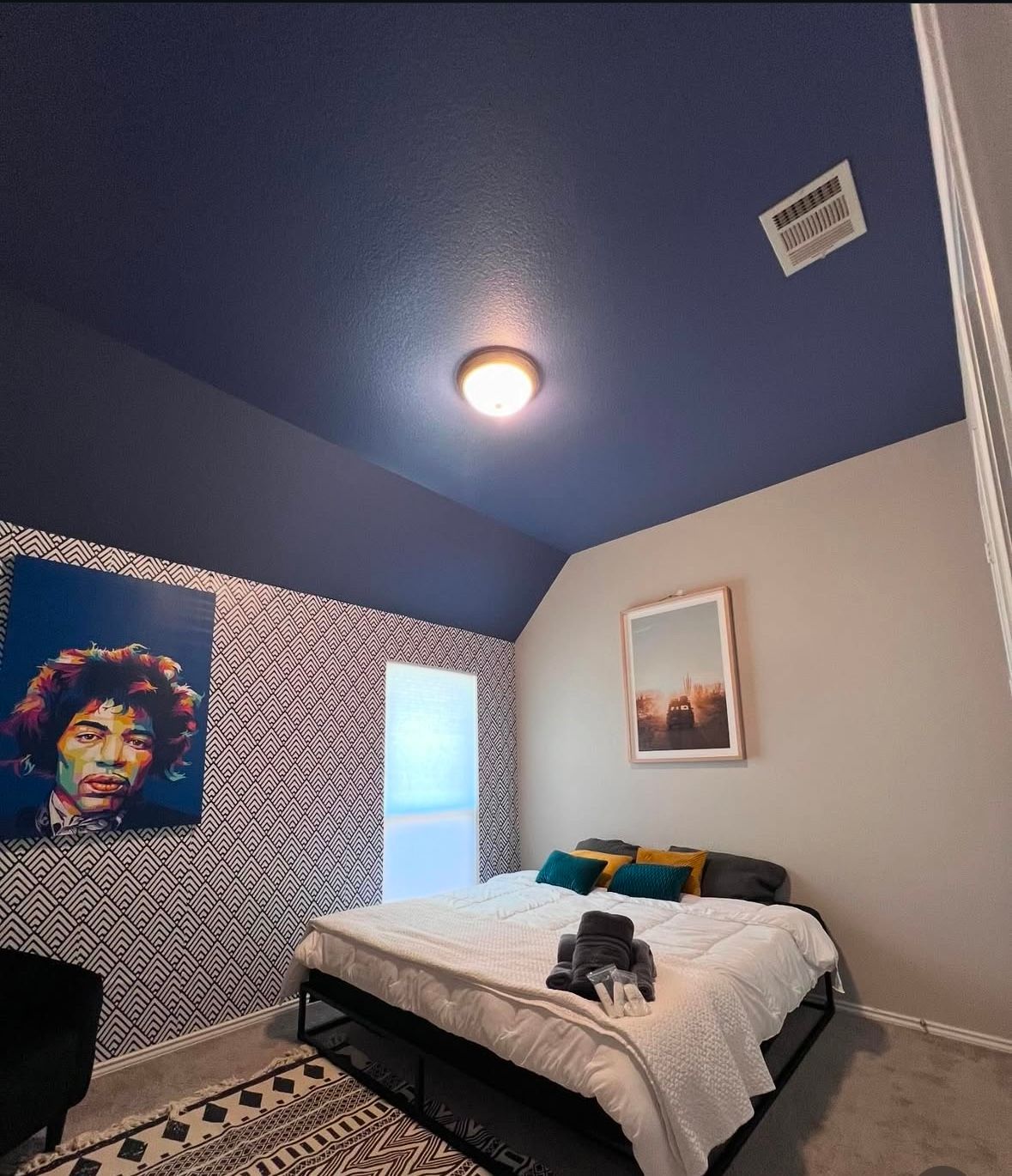 Small bedroom with blue ceiling, patterned wall, bed, colorful portrait, and window