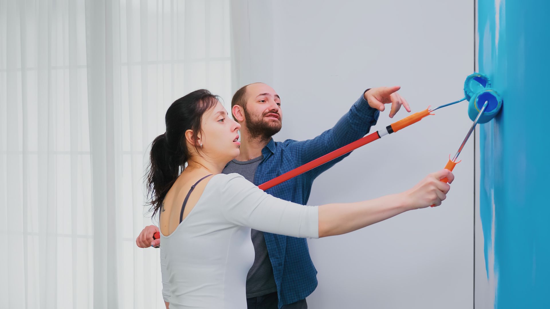 Couple painting a wall blue, with one pointing and the other rolling paint.