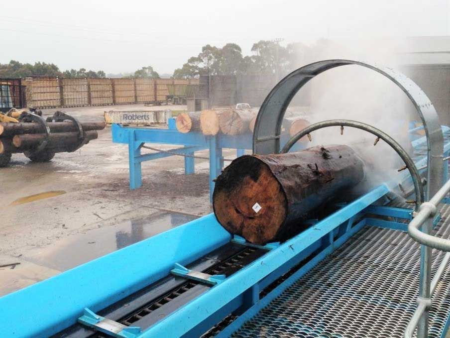 Log being washed by a water spray in a sawmill; blue machinery, industrial setting.