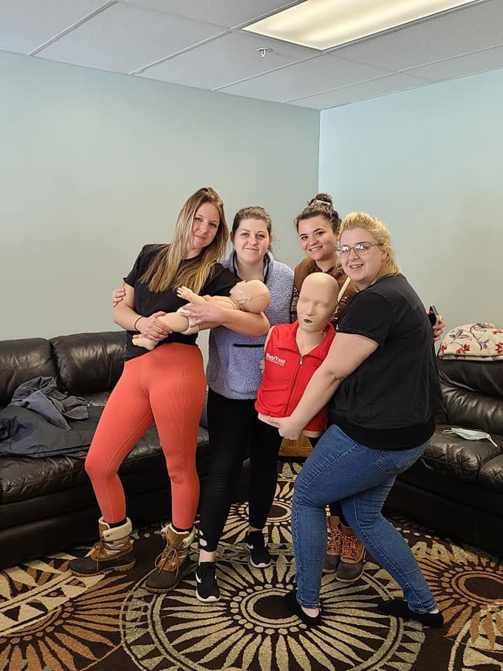 Group of women posing with CPR dummies. Some hold them. Indoors, neutral colors, and carpet.