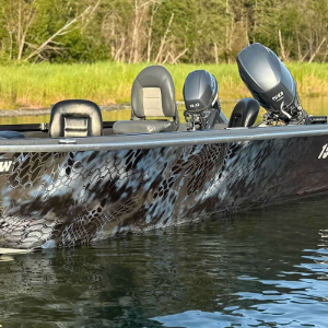 A camo-patterned aluminum fishing boat floats on a calm river with a grassy shoreline in the background.