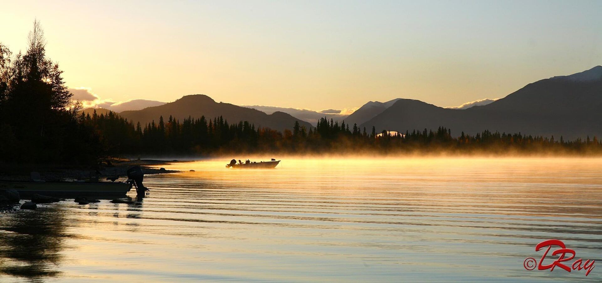 A small boat moves across a mist-covered lake at sunrise, with mountains in the background and a shoreline in shadow.