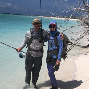 Two people in sun-protective clothing and backpacks stand together on a white sand beach by turquoise water, holding rods.