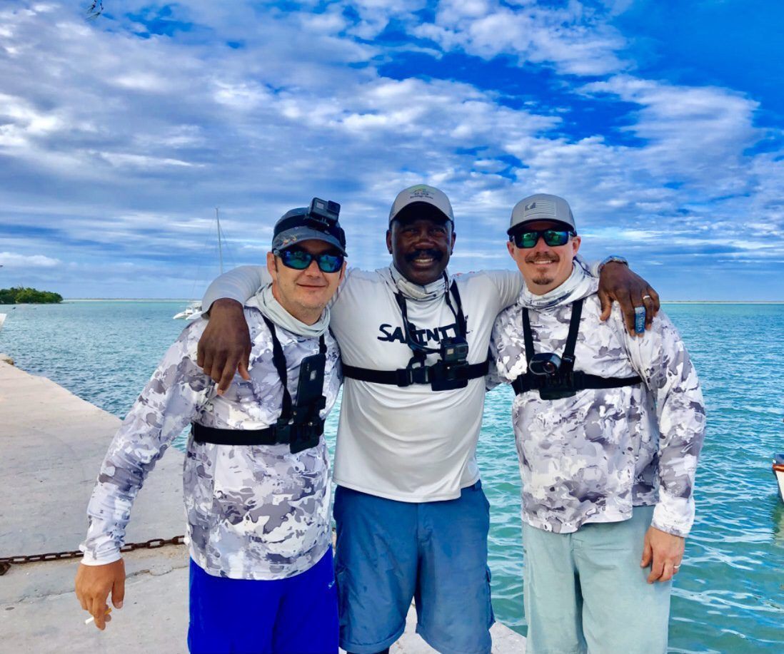Three people wearing fishing gear and chest harnesses smile while posing together on a pier by the ocean.