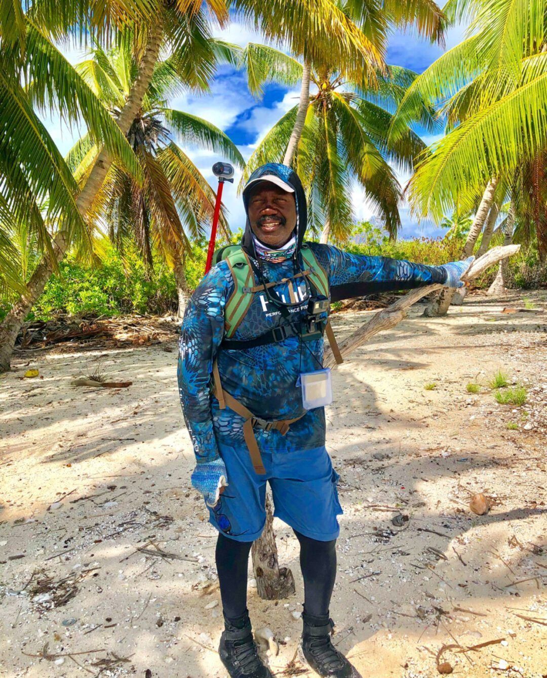 A person in blue camouflage gear smiles while standing on a sunny, palm-lined beach.